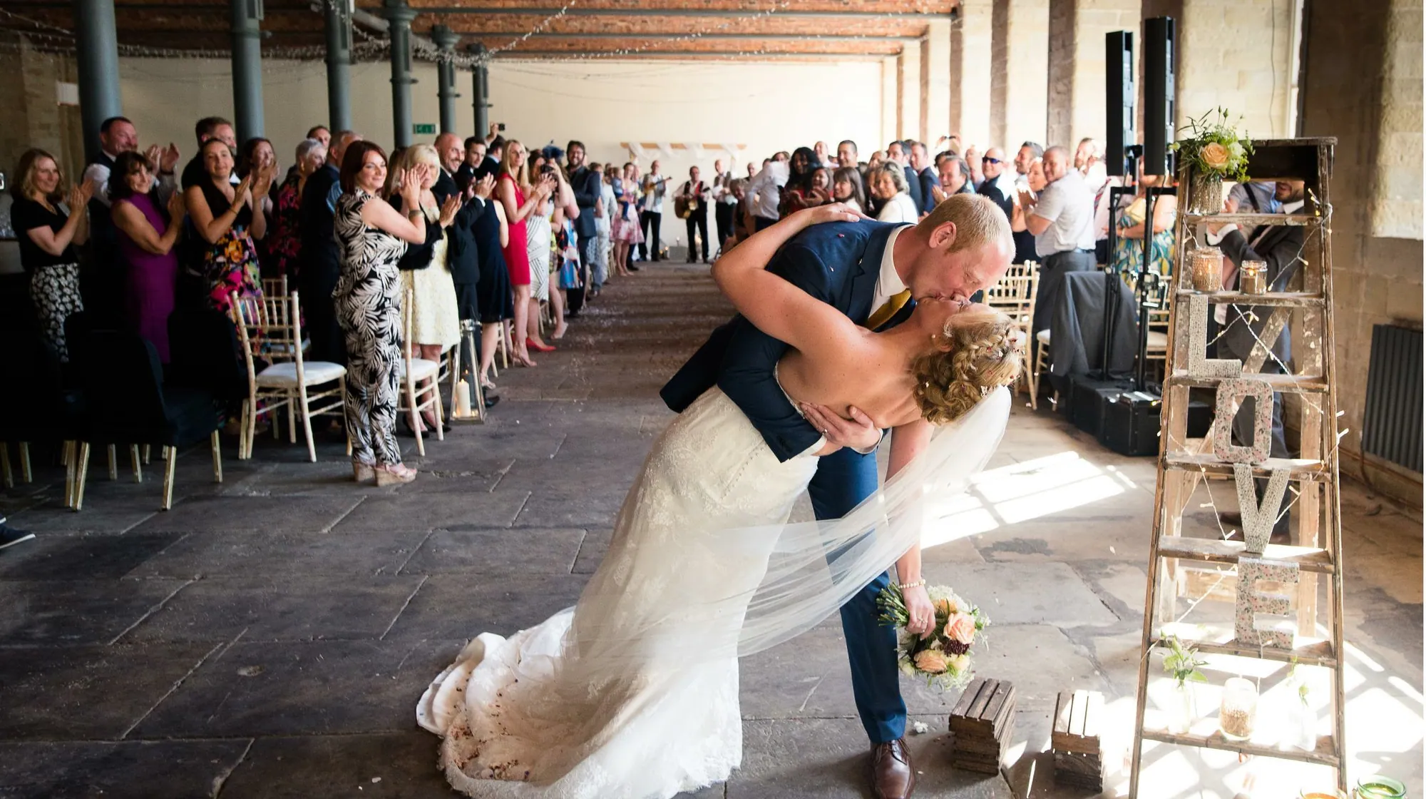 A couple kiss at the end of the aisle beneath the red-brick beams, guests applauding