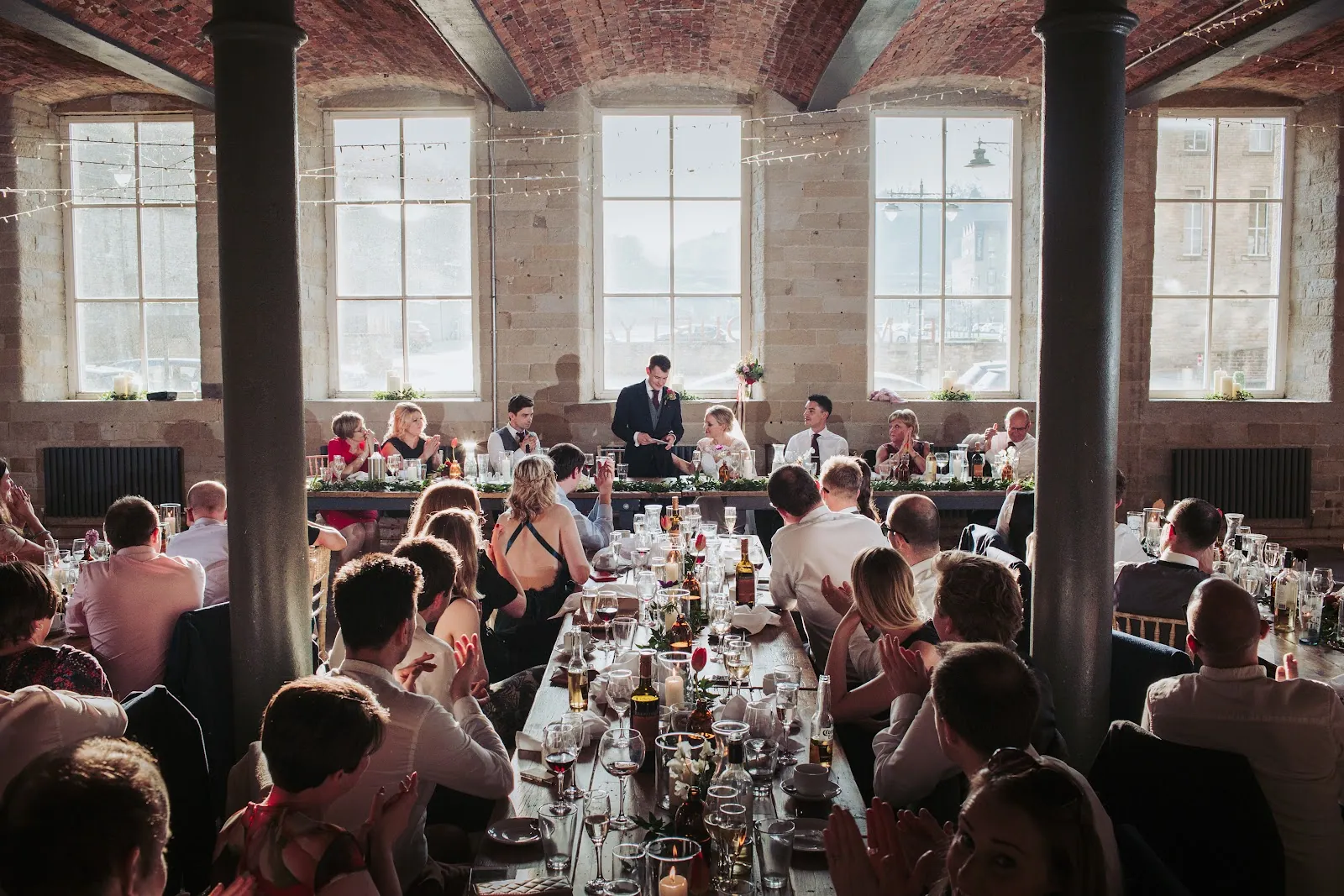 The grand hall at The Arches: a vaulted red-brick ceiling, cast-iron columns and tall arched windows above long tables of wedding guests